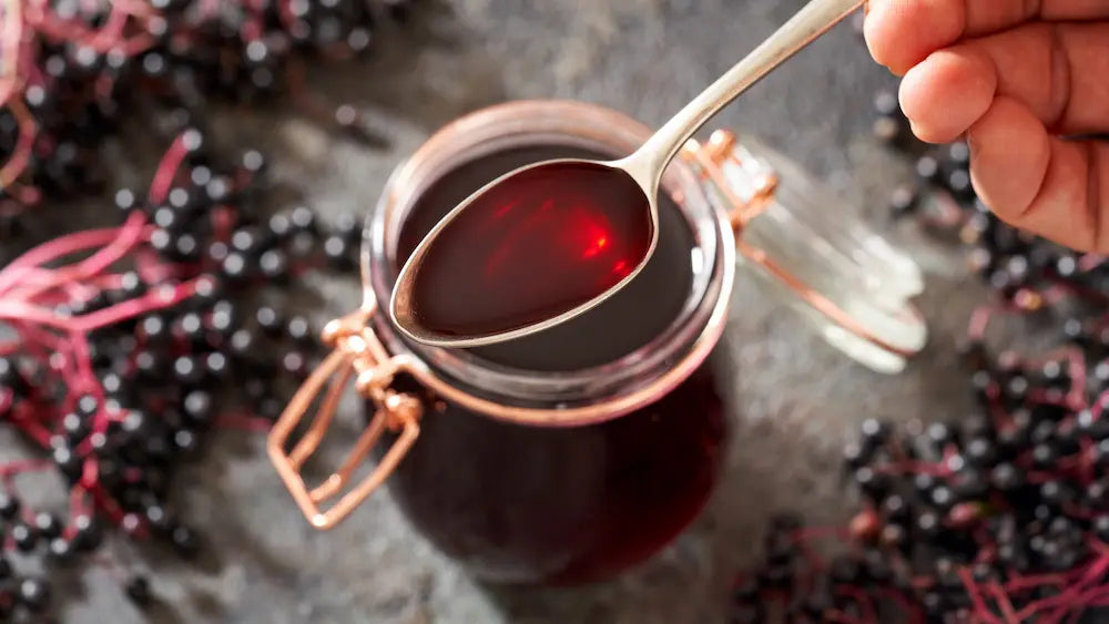 Measured spoon of deep-purple elderberry syrup beside black elderberries in a softly lit New Zealand kitchen