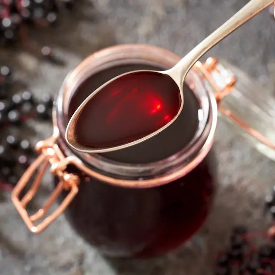 Measured spoon of deep-purple elderberry syrup beside black elderberries in a softly lit New Zealand kitchen