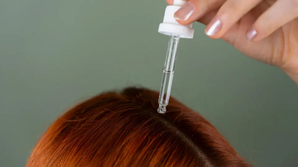 Adult gently parting intact hair near the scalp while holding an amber oil bottle in a bright bathroom