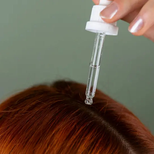 Adult gently parting intact hair near the scalp while holding an amber oil bottle in a bright bathroom